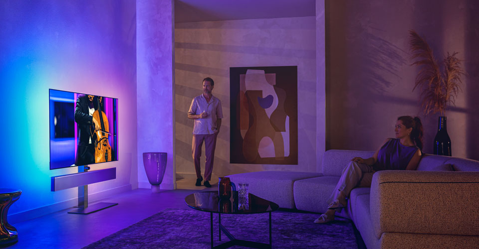 Bărbat și femeie &icirc;ntr-un living modern, urmărind un televizor Philips Ambilight care redă un spectacol de violoncel