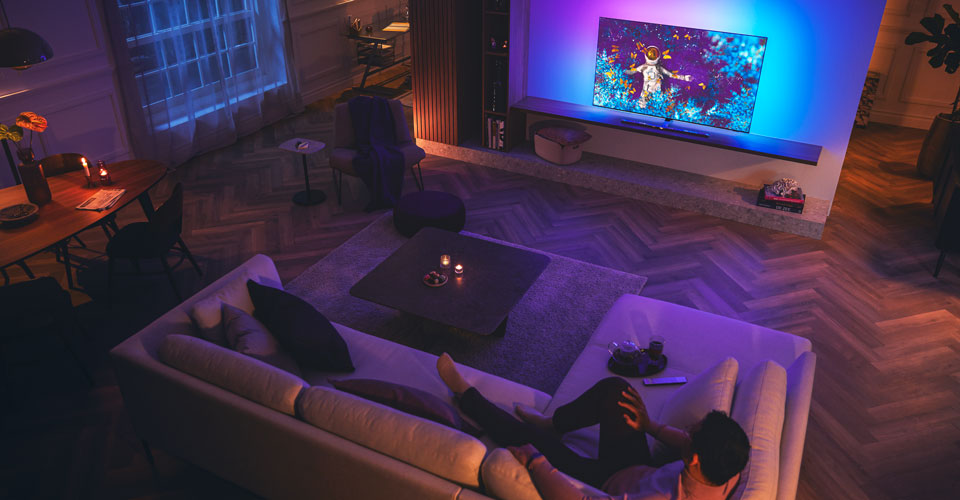 Bărbat relax&acirc;ndu-se pe o canapea &icirc;ntr-un living slab luminat, privind un televizor Philips Ambilight
