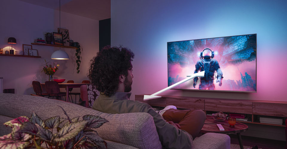 Bărbat așezat pe o canapea, urmărind un televizor Philips Ambilight care afișează o scenă colorată cu un astronaut, cu o săgeată care indică distanța optimă de vizionare